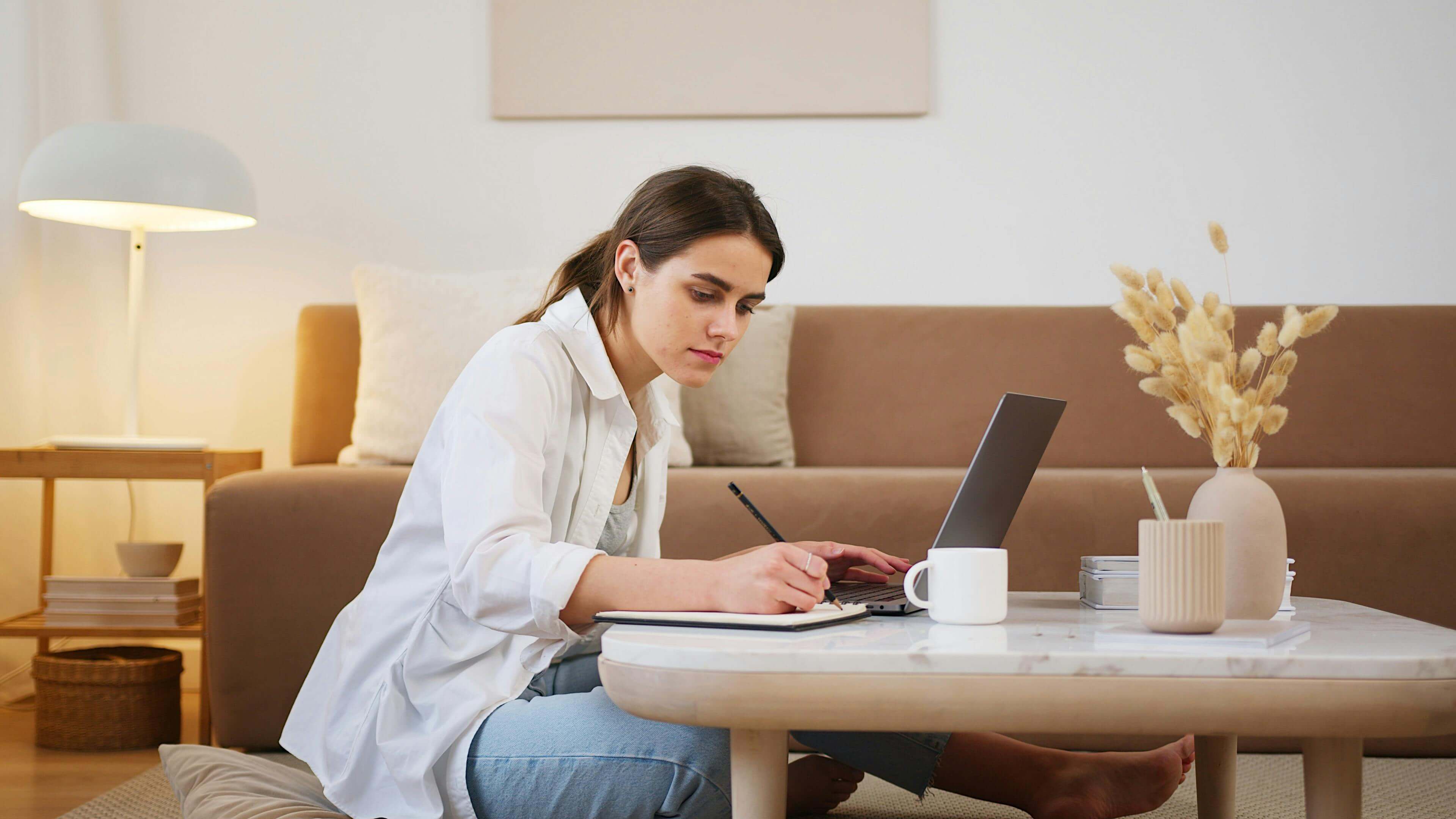Woman sitting on ground with low table in living room in front of computer taking notes.